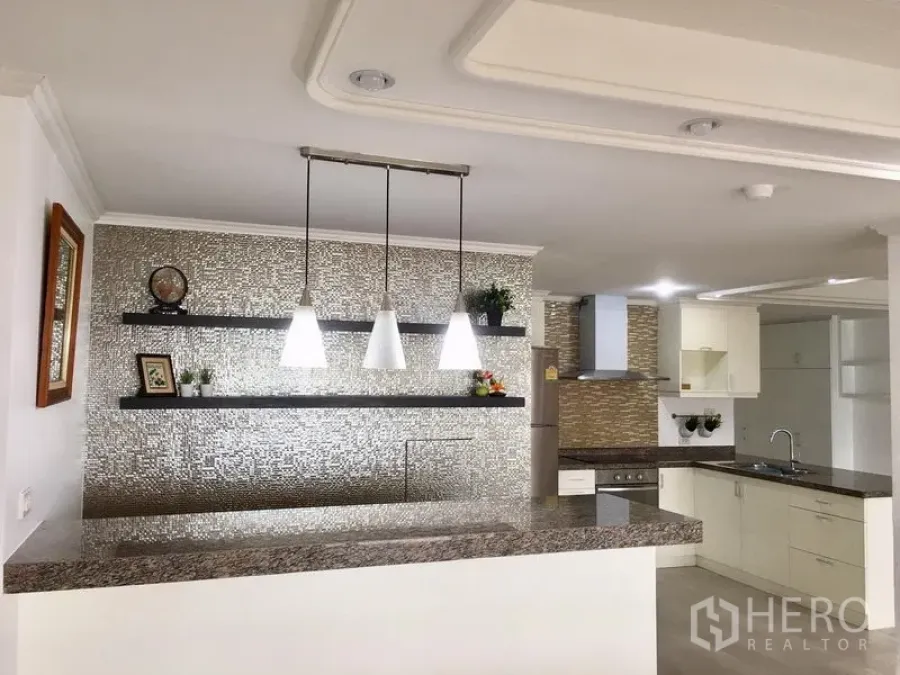 Condominium for sale in Watthana, Bangkok - Breakfast bar with pendant lights, floating shelves and textured accent wall beside an L-shaped kitchen with stone countertops.