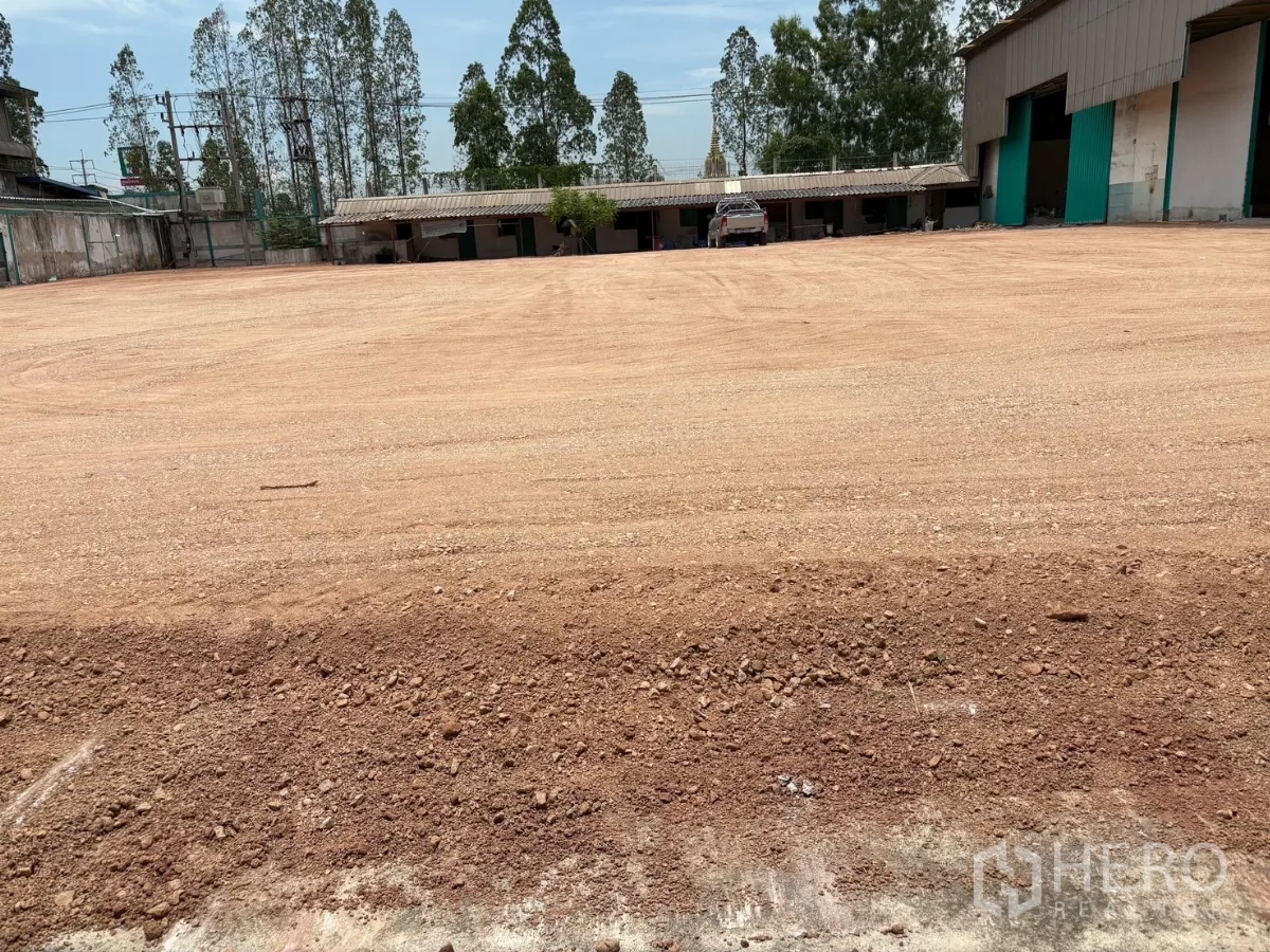Factory for rent in Mueang Samut Sakhon, Samut Sakhon - Expansive open yard of leveled red earth with boundary trees and site buildings.