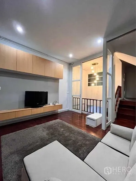 Townhouse for rent in Khlong Toei, Bangkok - Family lounge with TV unit, sofa, rug and glass partition by the stairs.