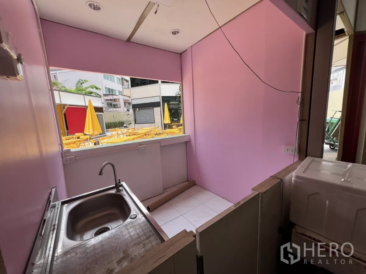 Retail Space for rent in Khlong Toei, Bangkok - Compact service counter with stainless sink and pink walls opening to a courtyard with yellow outdoor umbrellas and seating.