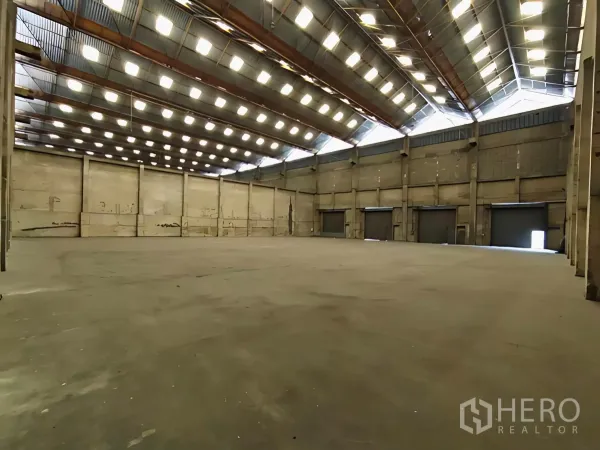 Factory for rent in Khlong Toei, Bangkok - Spacious factory interior with a high roof, skylights, concrete floor, and multiple roller shutter doors.