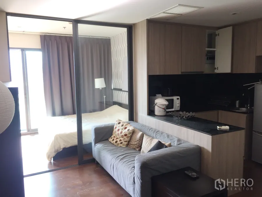 Condominium for rent in Khlong Toei, Bangkok - Open-plan living area with grey sofa, compact kitchen with black countertop, and a bedroom behind sliding glass.