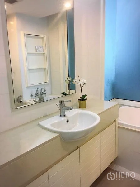 Condominium for rent in Khlong Toei, Bangkok - Bathroom vanity with large mirror, storage cabinets, and tub beside a window.