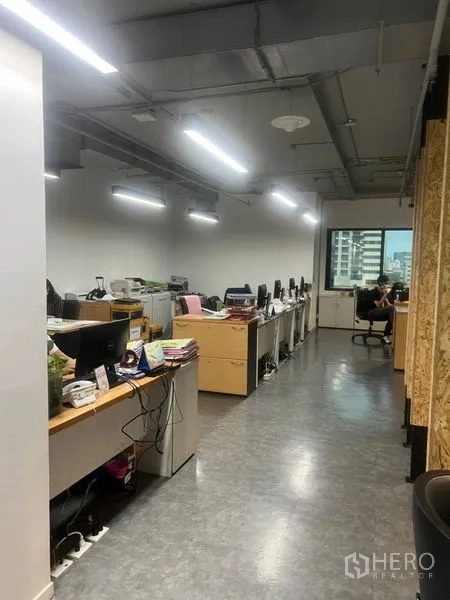Office Space for rent - Open-plan office corridor with long workstations, LED strip lighting, exposed ceiling, and a city-view window.