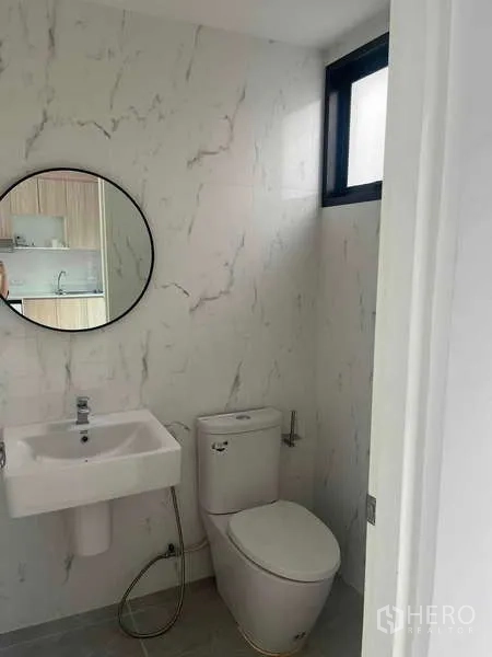 Townhouse for rent in Suan Luang, Bangkok - Compact bathroom with marble-effect walls, round mirror, square basin, toilet and a small window.