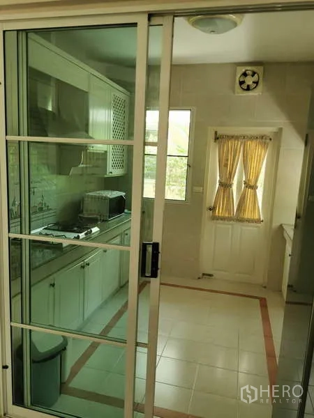 Detached House for rent - Separate enclosed kitchen behind a glass sliding door with white cabinets, cooktop, and a window with yellow curtains.