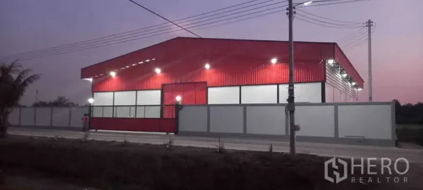 Warehouse for rent or sale in Khlong Luang, Pathum Thani - Dusk street-front view of the red warehouse facade and walled compound with exterior lights.