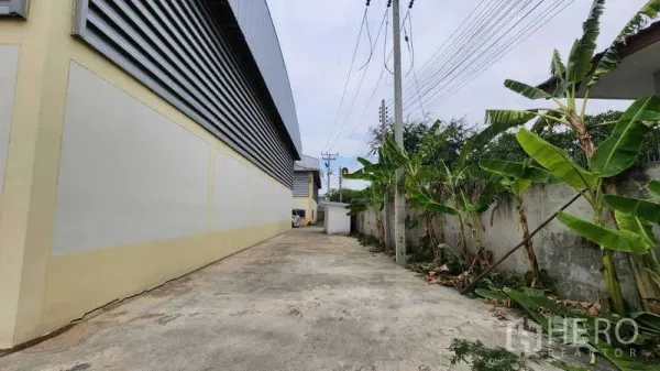 Factory for rent in Ban Phaeo, Samut Sakhon - Paved side driveway beside the factory with metal louvered walls, power poles and greenery for truck access.
