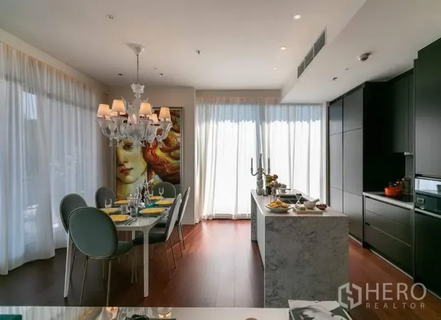 Condominium for sale in Watthana, Bangkok - Bright open-plan kitchen and dining area with a marble island, chandelier lighting, dark cabinetry, and floor-to-ceiling curtains.