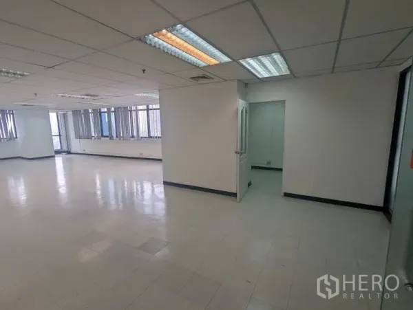 Office Space for rent or sale - Open-plan office with tiled floor, drop ceiling, fluorescent lights and a doorway to a small private room, plus long windows with blinds.