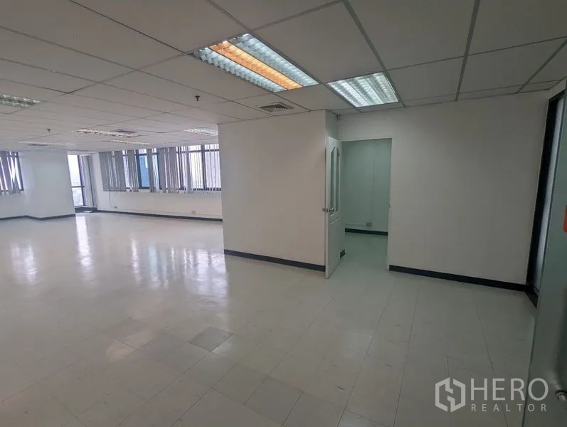 Office Space for rent or sale - Open-plan office with tiled floor, drop ceiling, fluorescent lights and a doorway to a small private room, plus long windows with blinds.