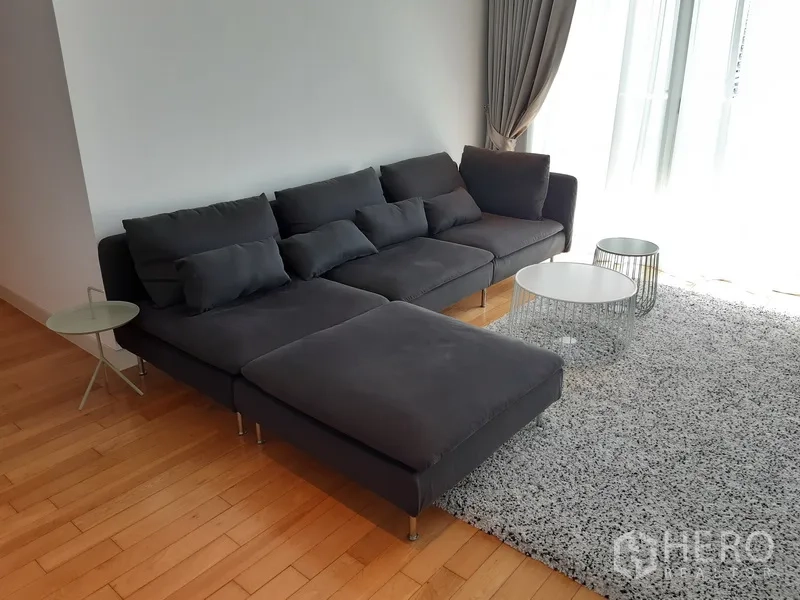 Condominium for rent in Khlong Toei, Bangkok - Bright living room with dark L-shaped sofa, shag rug and floor-to-ceiling curtains on hardwood floors.