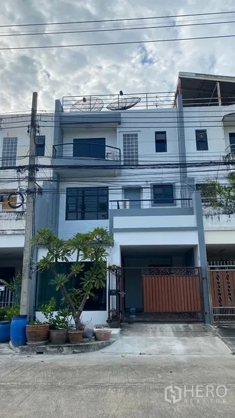 Townhouse for rent in Bang Na, Bangkok - Three-storey townhouse facade in Bang Na, Bangkok with gated carport and potted plants.