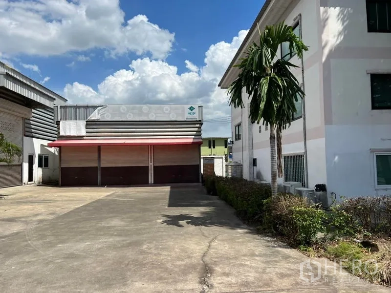 Warehouse for rent in Prawet, Bangkok - Front view of a warehouse with wide concrete yard, red canopy and multiple roller shutters beside an office building and palm tree under a blue sky.