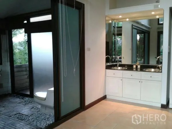 Detached House for rent in Suan Luang, Bangkok - Bathroom with twin sinks, large mirror, and a frosted-glass door to a semi-outdoor shower and toilet.