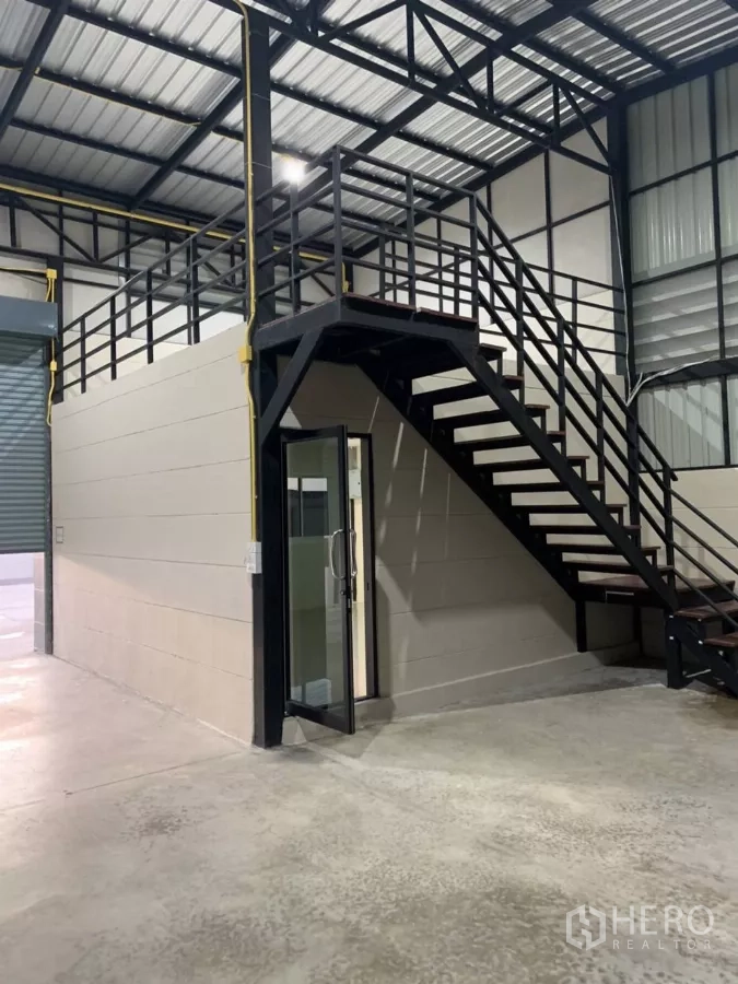Warehouse for rent in Bang Bua Thong, Nonthaburi - Warehouse interior showing a steel staircase to a mezzanine, glass-door office entry, high metal roof and concrete floor.