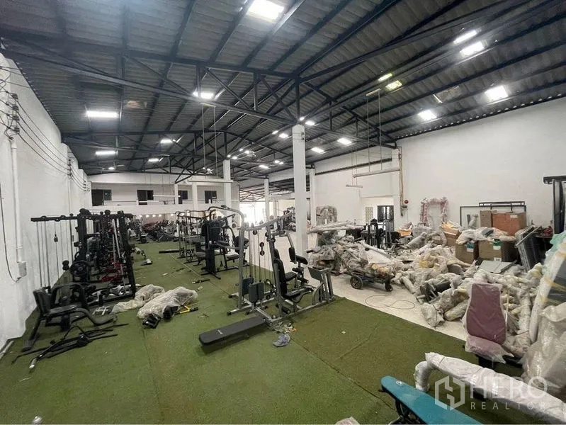 Warehouse for sale in Lam Luk Ka, Pathum Thani - Interior storage zone showing rows of packed gym equipment on artificial turf under high‑bay lights.