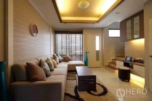 Townhouse for rent in Phra Khanong, Bangkok - Cozy living room with large sofa, cove lighting, blinds and stairs to upper floor.