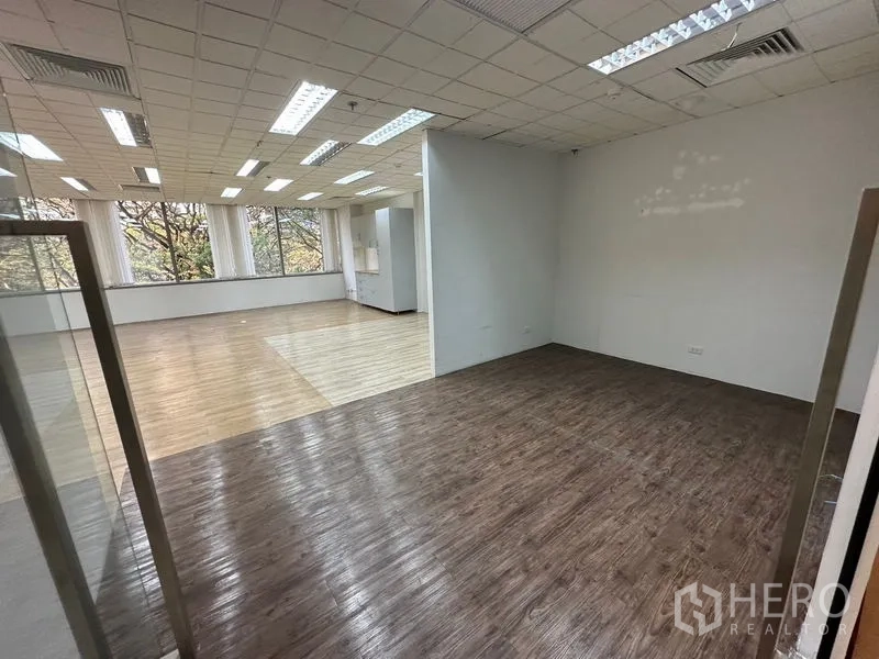 Office Space for rent - Glass-partitioned room with darker wood floor opening to a large office area with ceiling lights and tree views.