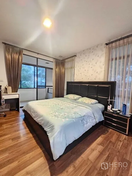 Detached House for sale in Prawet, Bangkok - Master bedroom with large bed, wood flooring, balcony windows and sheer curtains.