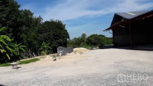 工厂 出售在Phanat Nikhom, Chon Buri - 金属屋顶厂房旁的大型碎石场地，周围树木茂盛，蓝天晴朗。