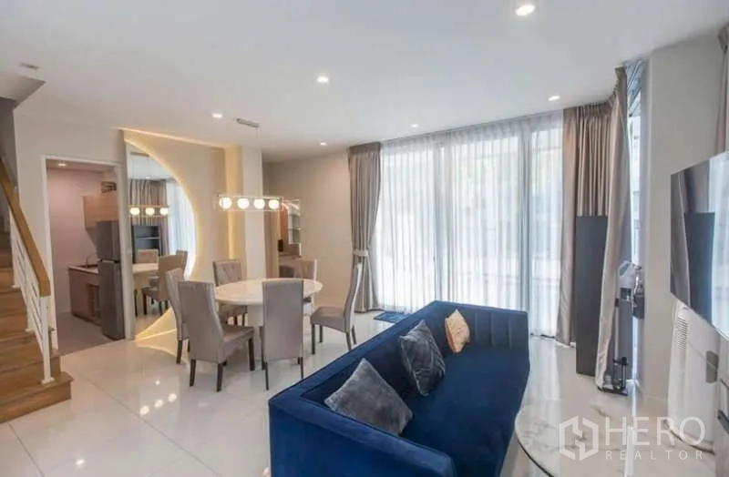 Detached House for rent or sale in Lat Phrao, Bangkok - Open-plan living and dining room with blue sofa, round table and full-height windows.