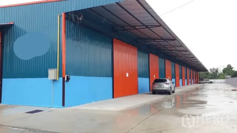 Factory for rent in Khlong Luang, Pathum Thani - Row of modern steel factory units with red sliding doors, covered loading canopy and wide concrete driveway in Khlong Luang.
