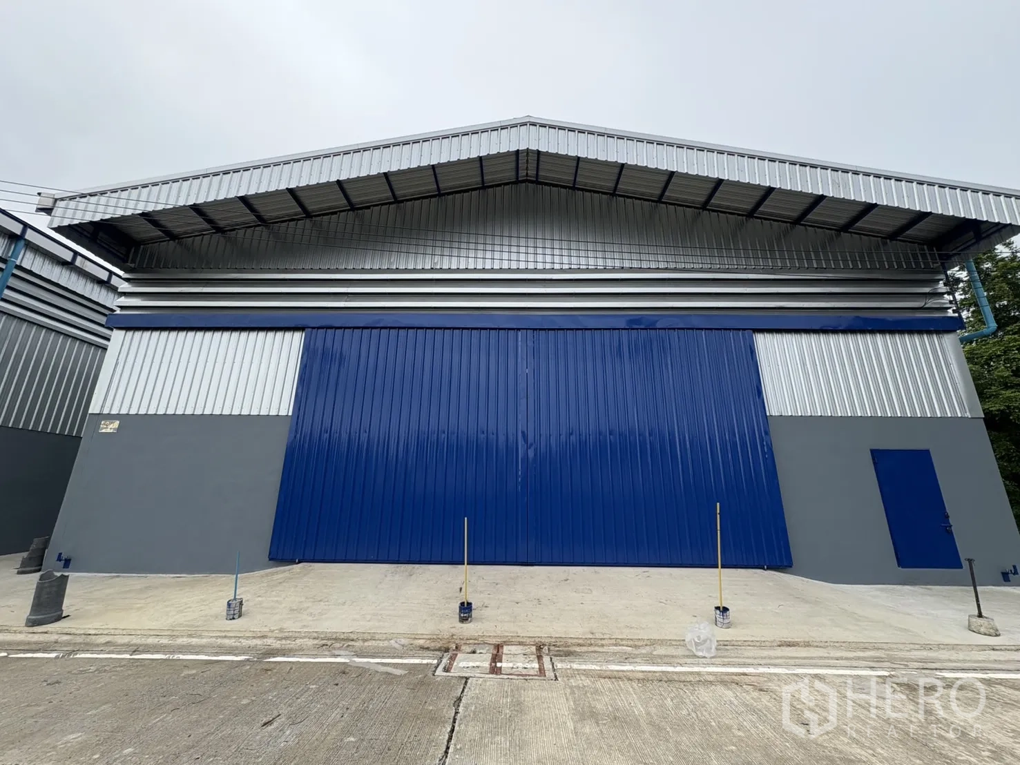 Warehouse for rent in Bang Phli, Samut Prakan - Front view of a metal-clad warehouse with large blue sliding doors and a wide concrete driveway.