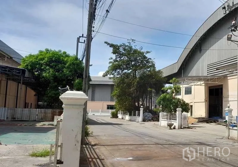 Factory for rent in Mueang Samut Prakan, Samut Prakan - Access road between factory buildings with trees, security fence and parking area.