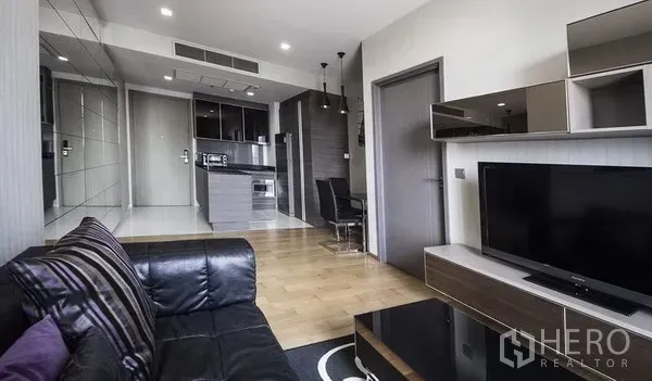 Condominium for rent in Khlong Toei, Bangkok - Open-plan living room with leather sofa, TV console, and compact kitchen with dining in a Sukhumvit condo.