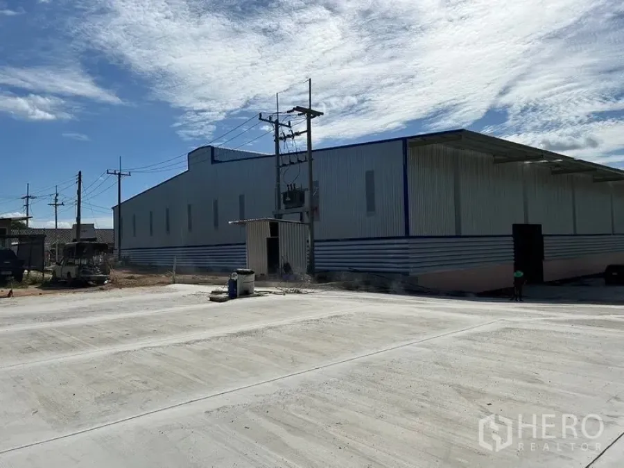 Warehouse for rent in Nikhom Phatthana, Rayong - Metal-clad warehouse exterior with large concrete yard and overhead power lines under a blue sky in Nikhom Phatthana.