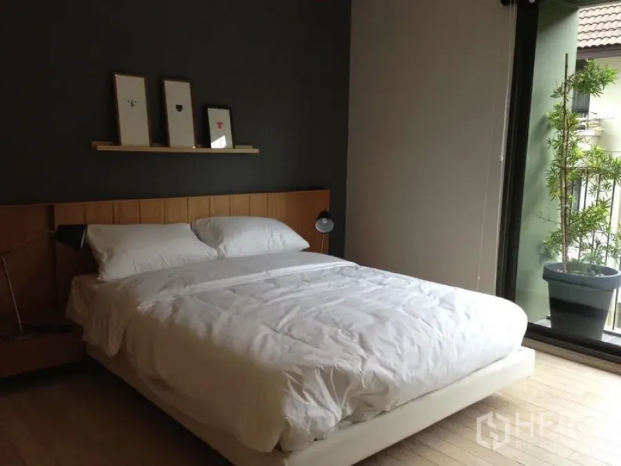 Condominium for rent in Watthana, Bangkok - Bright bedroom with double bed, dark accent wall, shelf with frames, and balcony with potted plant.