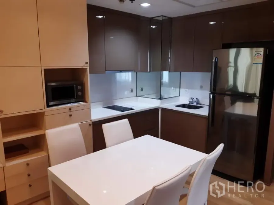 Condominium for rent in Khlong Toei, Bangkok - Modern U-shaped kitchen with glossy cabinets, cooktop, microwave, fridge, and a white four-seat dining table.