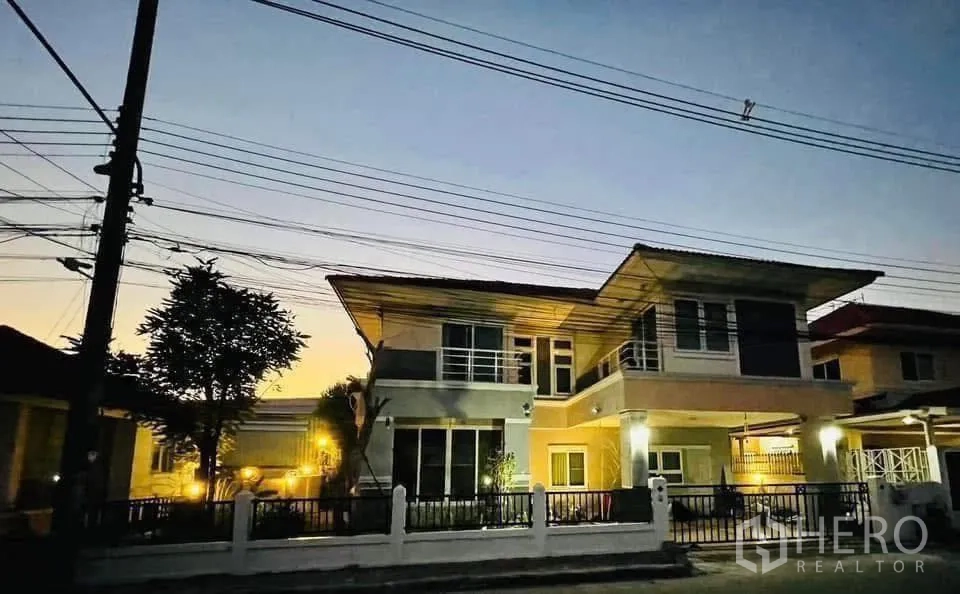 Detached House for rent or sale in Lam Luk Ka, Pathum Thani - Two-storey detached house with balcony, gated fence and warm exterior lights at dusk in Lam Luk Ka.