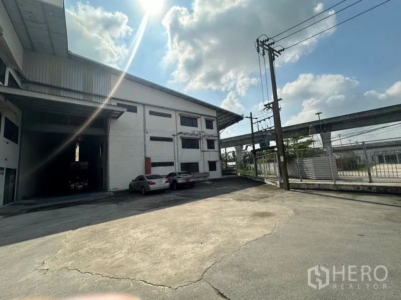 Warehouse for rent in Suan Luang, Bangkok - Front yard and gated entrance of the warehouse with power poles and parking area.
