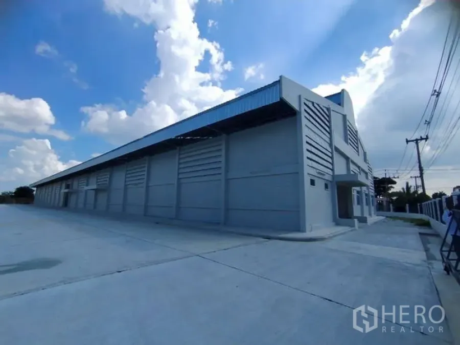 Warehouse for rent in Bang Pakong, Chachoengsao - Exterior of a large warehouse with expansive driveway and power lines along the road.