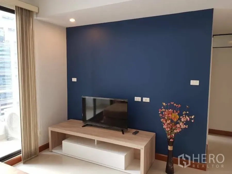 Condominium for rent in Watthana, Bangkok - Feature wall in navy blue with TV on a wooden media unit and a vase, next to balcony sliding door.