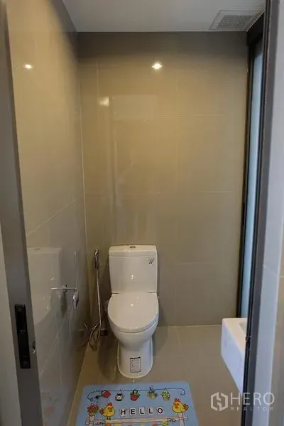 Condominium for rent in Khlong Toei, Bangkok - Compact bathroom with beige tiles, modern toilet and bidet spray beside a glass door.