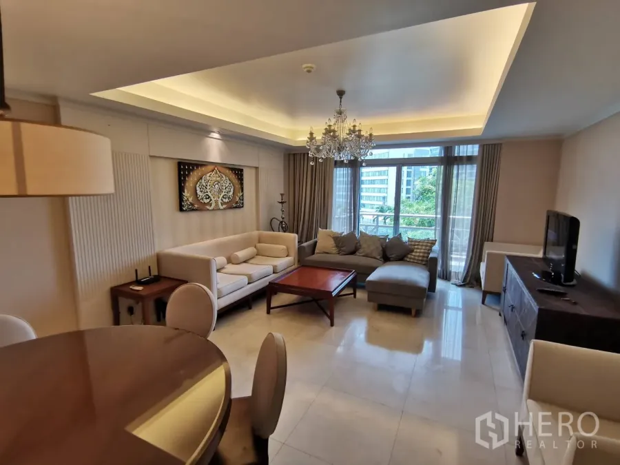 Condominium for rent in Pathum Wan, Bangkok - Bright living room with chandelier, L‑shaped sofas and sliding doors to a balcony overlooking greenery and city views.