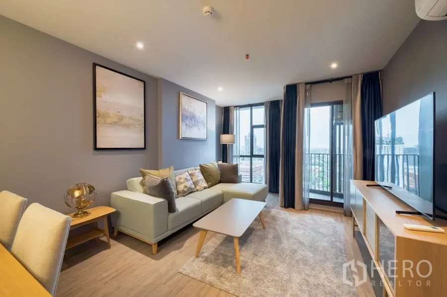 Condominium for rent in Khlong Toei, Bangkok - Bright living room with L-shaped sofa, TV console and floor-to-ceiling windows overlooking the city skyline.