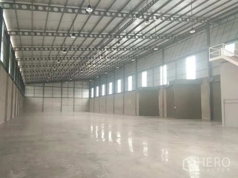 Warehouse for rent in Bang Phli, Samut Prakan - Expansive warehouse interior showing column-free space, high steel truss roof, windows for natural light, and multiple roller shutters.