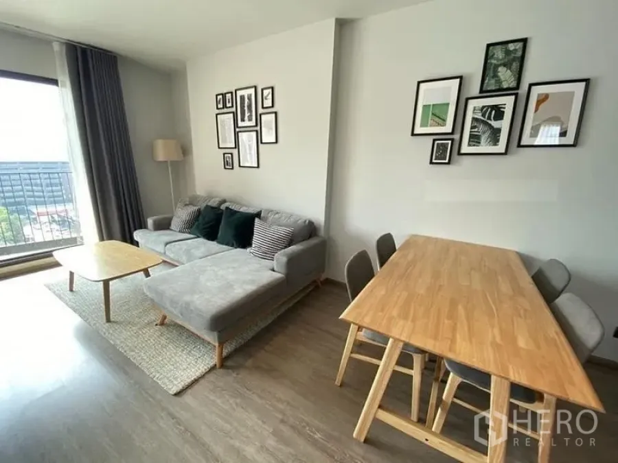 Condominium for rent in Watthana, Bangkok - Open-plan living and dining area with L-shaped sofa, wall art, and balcony access.