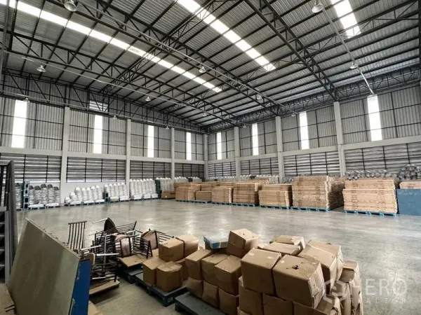 Warehouse for rent in Bang Phli, Samut Prakan - Open warehouse space with tall windows, ribbed metal walls and stacks of goods on pallets.