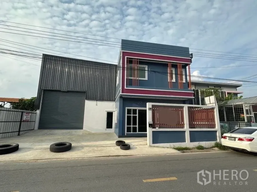 Warehouse for rent in Lam Luk Ka, Pathum Thani - Street-facing warehouse with large roller shutter beside a two-storey blue office, gated frontage and parking.