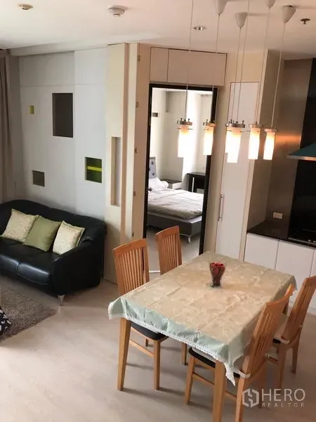 Condominium for rent in Khlong Toei, Bangkok - Open-plan dining area with four wooden chairs and pendant lights beside a compact kitchen, with a doorway leading to a bedroom.