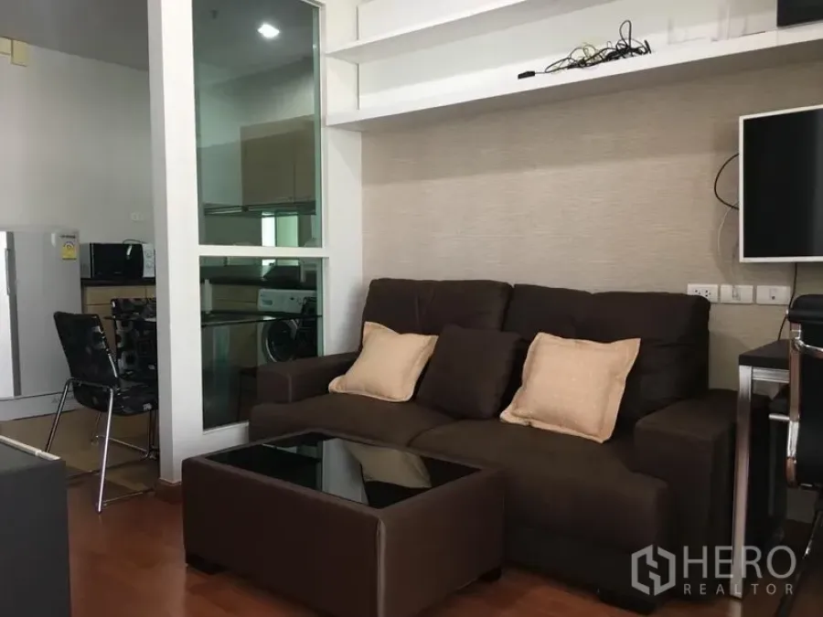 Condominium for rent in Pathum Wan, Bangkok - Cozy living room with brown sofa, glass coffee table, wall shelving and a window to the kitchen.