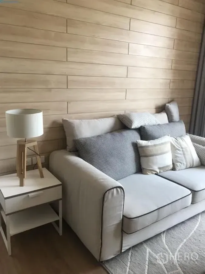 Condominium for rent in Watthana, Bangkok - Bright living room with beige sofa, side table and wooden lamp against a light wood panel wall.
