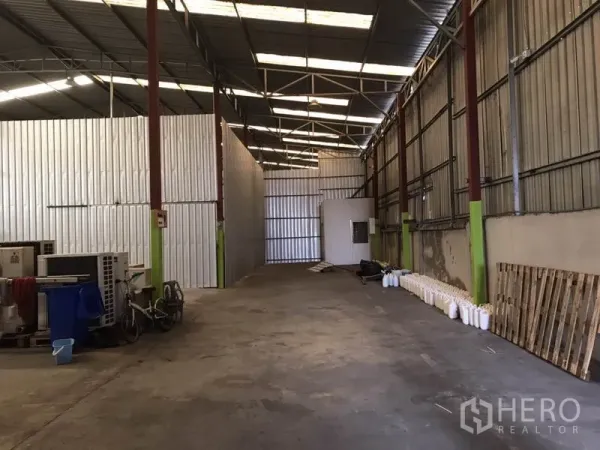 Warehouse for rent in Bang Na, Bangkok - Long aisle of the warehouse showing metal partitions, a small utility room and exposed roof trusses over a concrete floor.