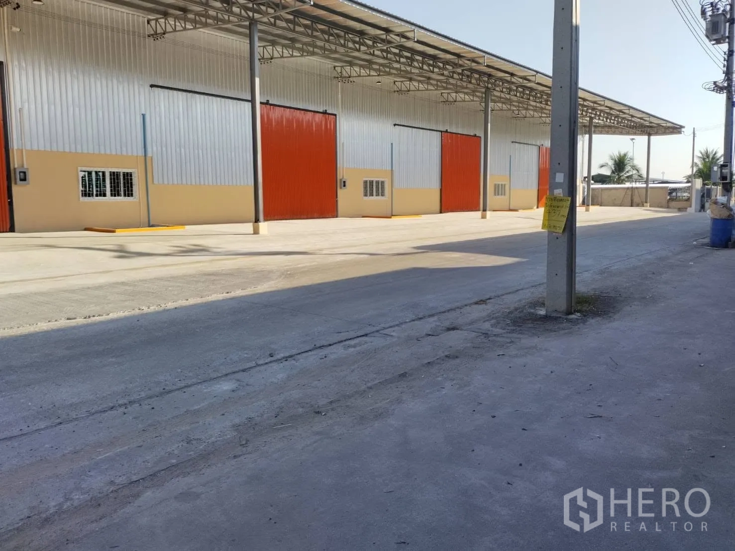 Warehouse for rent in Khlong Luang, Pathum Thani - Warehouse exterior with covered loading bay, multiple large sliding metal doors, and a spacious concrete yard along the access road.