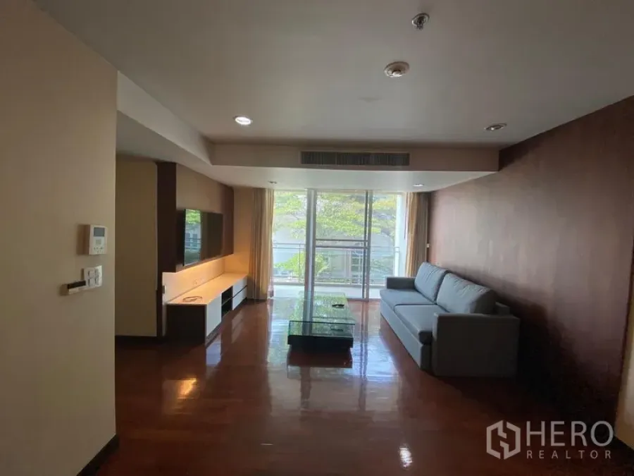 Condominium for rent in Watthana, Bangkok - Spacious living room with sofa, TV wall, wood floors and sliding glass doors to a green-view balcony.